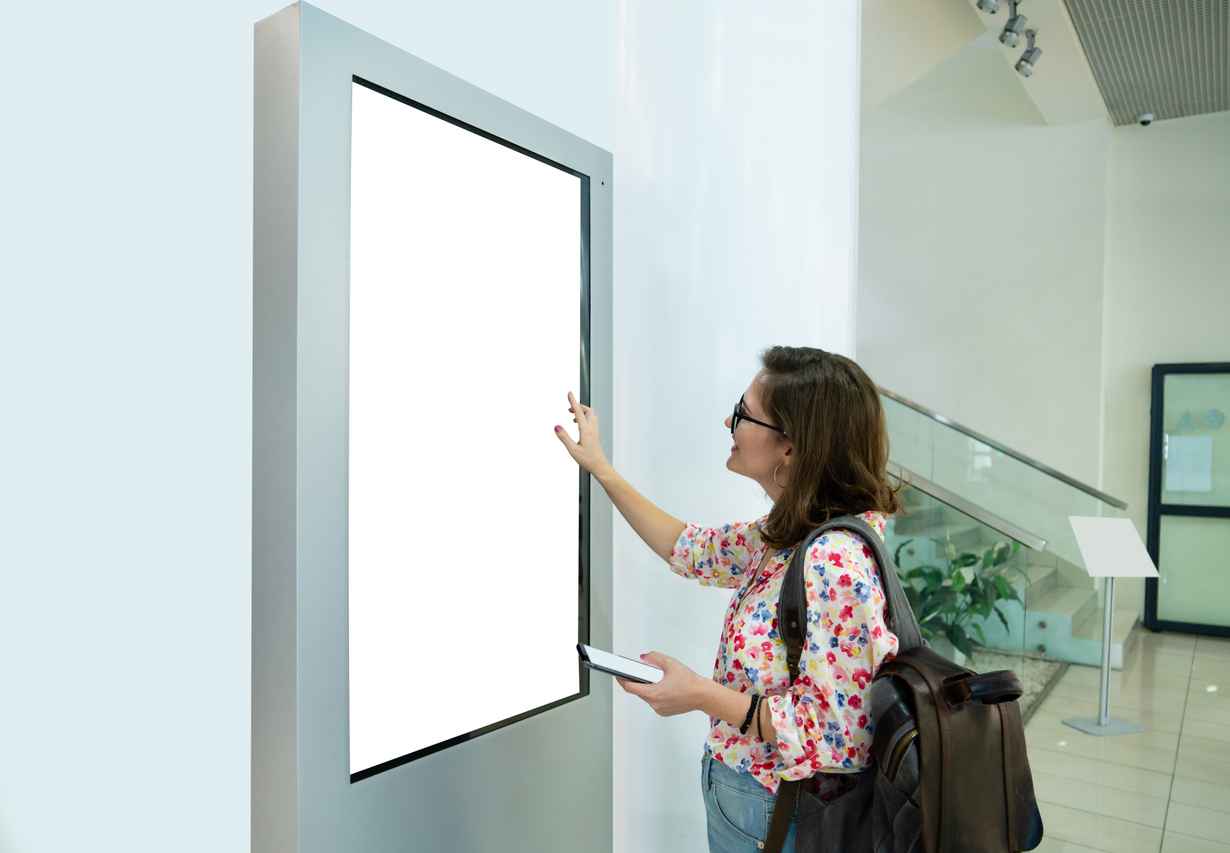 Woman with phone uses self-service desk with touch screen; digital signage